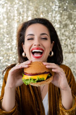 Altın küpeli gülen kadın parlak gümüş arka planda lezzetli bir burger tutuyor.