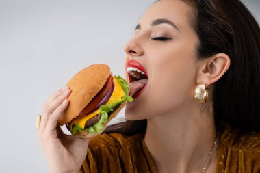 Makyajlı zarif genç bir kadın Gri üzerine izole edilmiş lezzetli bir burger yiyor.