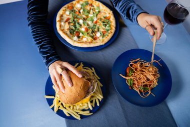 Mavi masada servis edilen patates kızartması ve pizzanın yanında hamburger ve makarna yiyen kadının kısmi görüntüsü