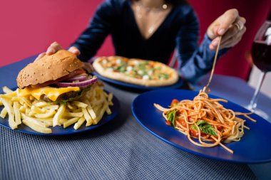 Burgerin seçici odağı patates kızartması ve pizzanın yanında spagetti ve kırmızı arka planı bulanık kesilmiş bir kadın.