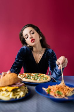 Seksi ve zarif bir kadın spagettiyi yanında pizza ve hamburgerle birlikte kırmızı masanın üzerine koyuyor.
