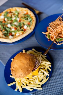Tabakların üst görüntüsü, spagetti ve patates kızartmasının yanında bulanık pizza ve mavi masada hamburger.