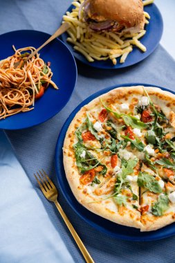 Makarna ve hamburgerin yanında leziz pizza ve mavi masada patates kızartması.