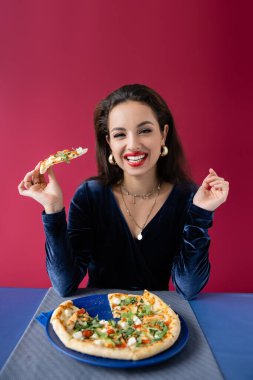 Elinde pizza tutan kadife elbiseli neşeli bir kadın ve kırmızı masada mavi masanın yanındaki kameraya bakıyor.