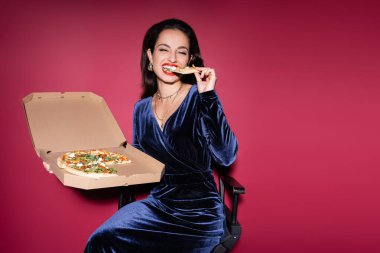 Mavi kadife elbiseli gülümseyen kadın kırmızı arka planda otururken lezzetli pizzalar yiyor.