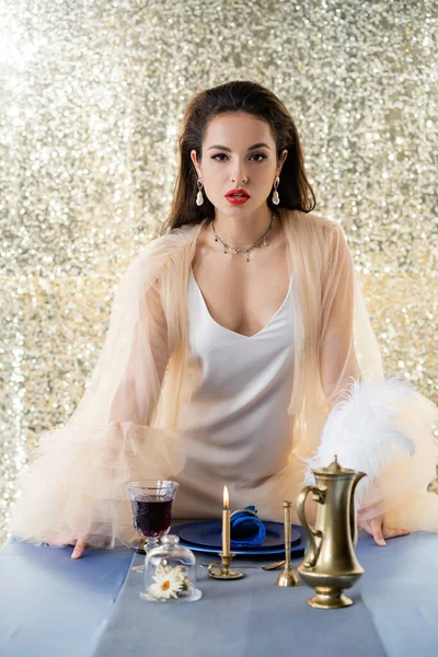 ブループレートの近くのカメラとキラキラした銀の背景のワインのガラスを見てエレガントなドレスの若い女性 — ストック写真