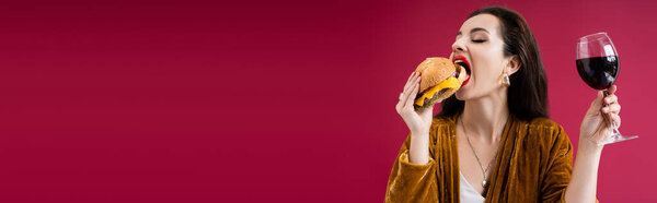 brunette woman with glass of wine eating delicious burger isolated on red, banner