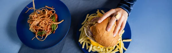 ブループレートの上でフレンチフライとスパゲティの近くでハンバーガーを食べているクロップドウィメンの — ストック写真