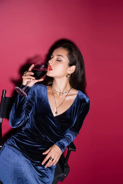 穿着蓝色天鹅绒衣服的时髦黑发女人 紧闭双眼 喝着红色背景的葡萄酒 — 图库照片