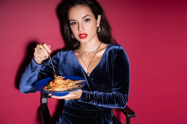sensual woman in elegant dress holding blue plate with pasta and looking at camera on red background