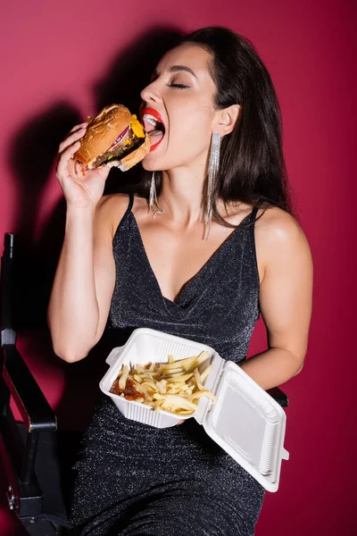 elegant woman eating tasty burger and holding plastic container with french fries on red background