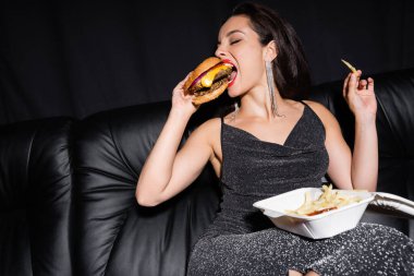 Lurex elbiseli genç bir kadın plastik konteynırın yanında hamburger yiyor ve patates kızartması siyahların üzerine izole edilmiş.