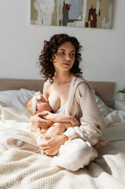 Kıvırcık anne yatakta oturuyor ve küçük kızı fiyonkla emziriyor. 