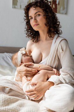 Kıvırcık saçlı kadın yatakta oturuyor ve saç bandıyla bebek emziriyor. 