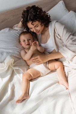 Yüksek açılı, kıvırcık elbiseli bir anne bebek kızıyla yatakta yatıyor. 
