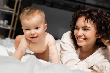 Kıvırcık saçlı neşeli anne yatakta emekleyen küçük kızına bakıyor.