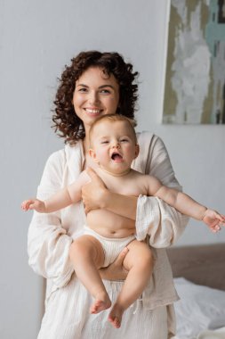 Pijamalı, gülümseyen bir kadın, küçük kızını yatak odasında tutuyor. 