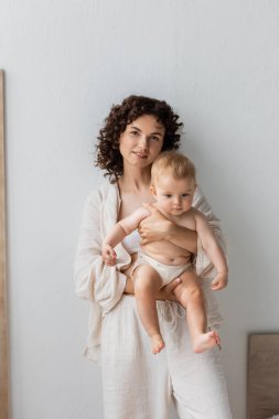 Pijamalı esmer anne kucağında küçük kızıyla evde kameraya bakıyor. 