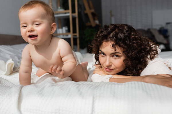 woman with curly hair looking at camera near curious baby daughter crawling on bed
