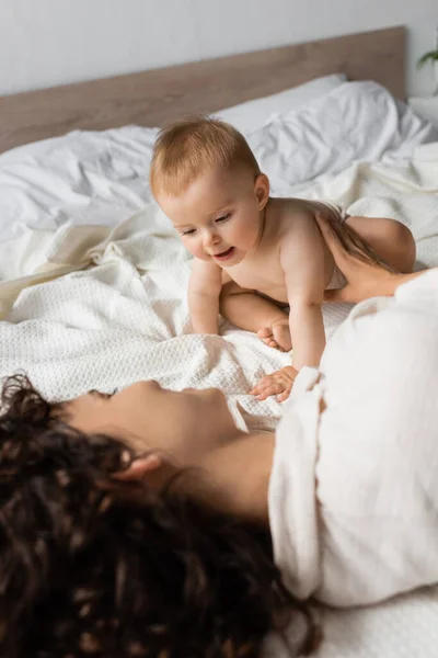 Säugling Sitzt Neben Fröhlicher Und Lockiger Mutter Auf Dem Bett — Stockfoto