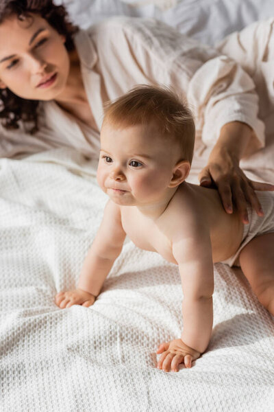 curly mother touching back of infant daughter crawling on bed