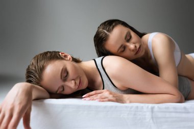 Gri arka planda gözleri kapalı bir çift seksi lezbiyen yatakta yatıyor.