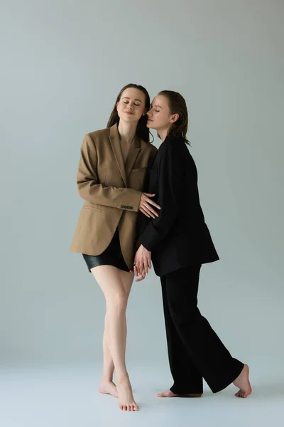 full length of barefoot lesbian girlfriends in blazers posing with closed eyes on grey background