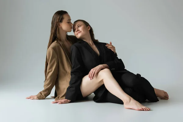 barefoot lesbian women in blazers smiling at each other while sitting on grey background