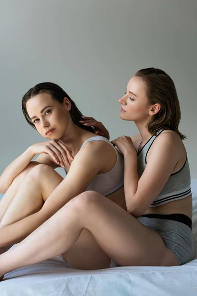 若い女性がベッドの上に座ってグレーで隔離されたカメラを見ているレズビアンのガールフレンドの髪に触れる — ストック写真