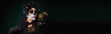 Santa Muerte kostümlü bir kadın koyu yeşil arka planda kafatası tutuyor. 