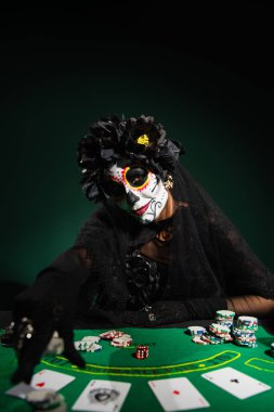 KYIV, UKRAINE - SEPTEMBER 12, 2022: Woman in mexican day of dead costume near playing cards on dark green  clipart