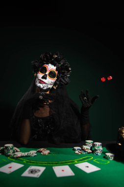 KYIV, UKRAINE - SEPTEMBER 12, 2022: Woman in halloween costume throwing dice near playing cards and chips on dark green  clipart
