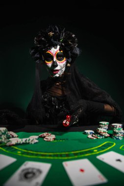 KYIV, UKRAINE - SEPTEMBER 12, 2022: Woman in day of death costume throwing dice near playing cards and chips on dark green background  clipart