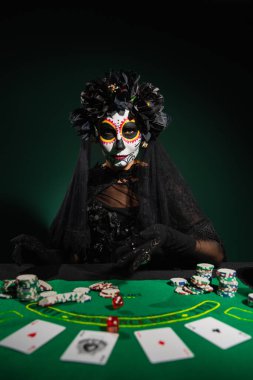 KYIV, UKRAINE - SEPTEMBER 12, 2022: Woman in mexican day of dead costume throwing dice on table on dark green clipart