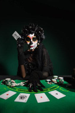 KYIV, UKRAINE - SEPTEMBER 12, 2022: Woman with sugar skull makeup and costume holding playing card on dark green clipart