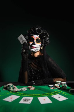KYIV, UKRAINE - SEPTEMBER 12, 2022: Woman in santa muerte costume holding playing card near chips on table on dark green  clipart