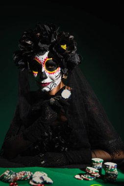 Woman in santa muerte halloween costume holding playing chip on dark green background  clipart