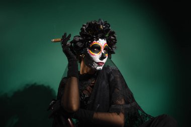Santa Muerte cadılar bayramı kostümlü bir kadın yeşil arka planda puro tutuyor. 