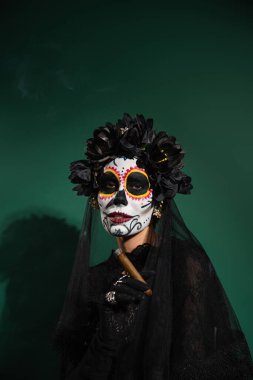 Santa Muerte Cadılar Bayramı kostümü ve yeşil arka planda puro tutan çelenkli kadın. 