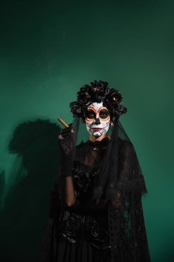 Santa Muerte cadılar bayramı kostümlü bir kadın yeşil arka planda puro tutuyor. 
