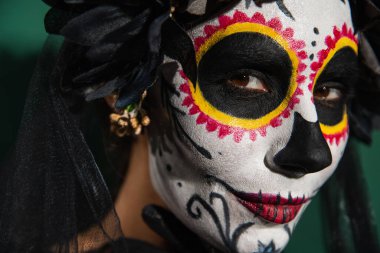 Santa Muerte makyajlı ve siyah peçeli bir kadının portresi yeşil arka planda kameraya bakıyor. 
