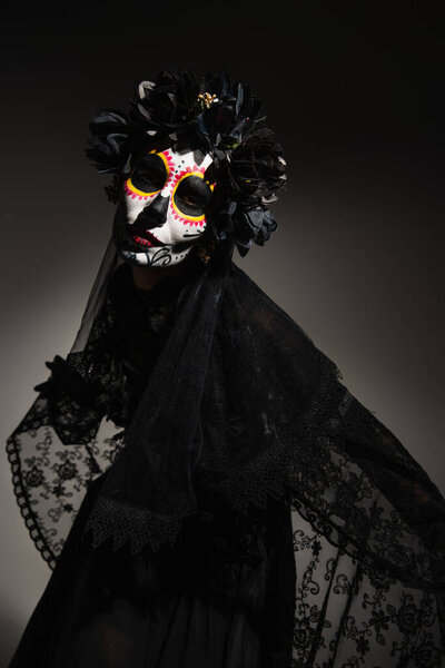 Woman with catrina makeup and dark wreath on head looking at camera on black background 