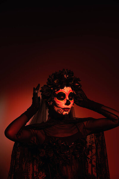 Woman in santa muerte traditional costume adjusting wreath on burgundy background with red lighting 