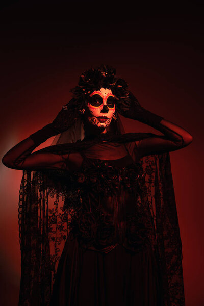 Woman in black costume and santa muerte makeup touching black wreath on burgundy background with red lighting