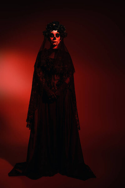 Full length of woman in mexican santa muerte costume looking at camera on burgundy background with red lighting