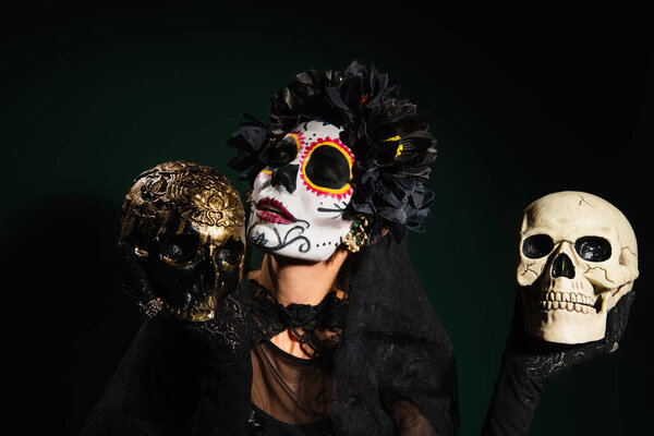 Woman in mexican santa muerte costume holding skulls on dark green background 