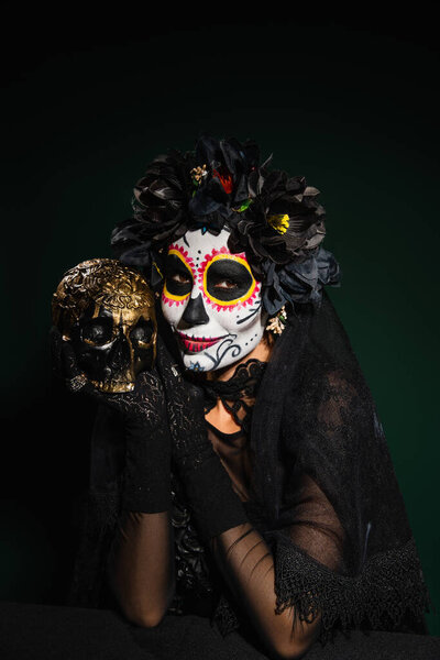 Smiling woman in mexican halloween costume holding skull on dark green background 