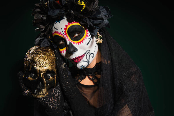 Woman in mexican catrina makeup and black wreath holding skull on dark green background 