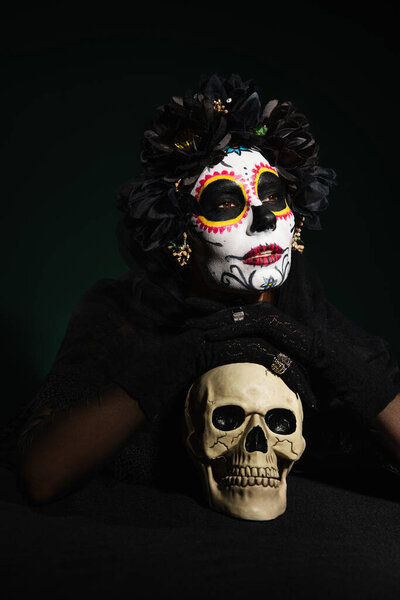 Woman in mexican day of dead makeup holding skull on dark green background 