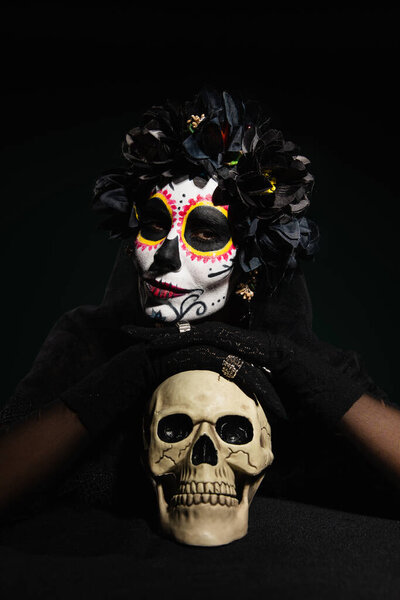 Женщина в мексиканском костюме в день смерти смотрит в камеру рядом с черепом, изолированным на черном 
